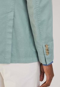 Giacca verde chiaro con un design strutturato, caratterizzata da una fila singola di bottoni marroni sui polsini e dettagli blu sul bordo delle maniche.