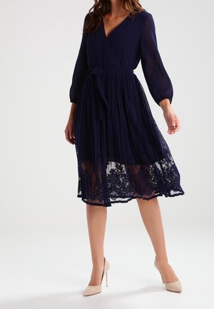 Frau trägt ein marineblaues knielanges Kleid mit transparenten langen Ärmeln, einem plissierten Rock, Spitzenbesatz am Saum und beigefarbenen High-Heels.