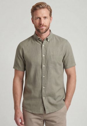 Man met een lichte baard die een olijfgroen kortemouw overhemd met knopen en een beige broek draagt, staat tegen een effen lichte achtergrond.