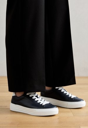 Czarne niskie sneakersy z białymi sznurowadłami i grubymi białymi podeszwami, noszone z czarnymi spodniami o szerokich nogawkach na drewnianej podłodze.