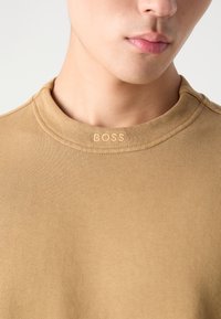 Sudadera de cuello redondo en color beige, hecha de un tejido acanalado; cuenta con un logotipo en relieve de "BOSS" en el cuello; textura suave con ligero estiramiento.