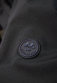 Svart tygpatch med Lindbergh-logotypen, omgiven av en cirkulär kant, med upphöjda bokstäver och designelement.