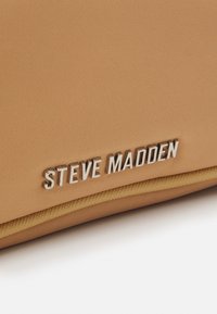Borsa di colore beige con texture, caratterizzata da un logo metallico argento "STEVE MADDEN" e un accento in oro lungo la cucitura.