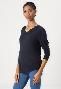 Pull marin avec un décolleté en V bordé de dentelle, des manches longues et une coupe décontractée. Associé à un jean bleu, mettant en avant des textures lisses.