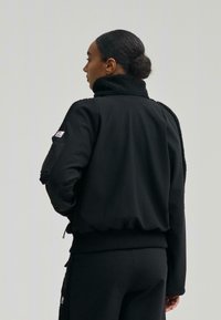 Černá bomberová bunda se měkkým, huňatým límcem, vroubkovanými manžetami a spodním lemem. Obsahuje boční zipovou kapsu a minimalistický design.