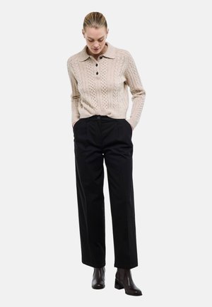 Femme portant un pull beige à torsades, un pantalon noir taille haute à jambes larges et des bottines noires, debout les mains dans les poches.