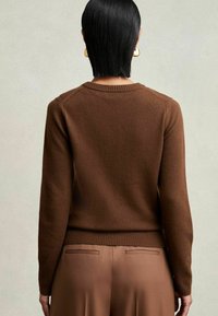Modelo de espaldas, vistiendo un suéter marrón y pantalones de tiro alto marrones. Su cabello negro liso y recto resalta un estilo minimalista.