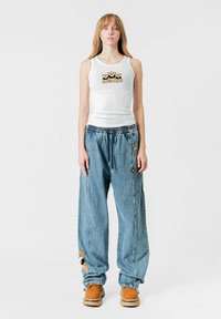 Camiseta sin mangas blanca con diseño de oso, combinada con pantalones de mezclilla sueltos de color azul claro con acentos bordados y calzado marrón.