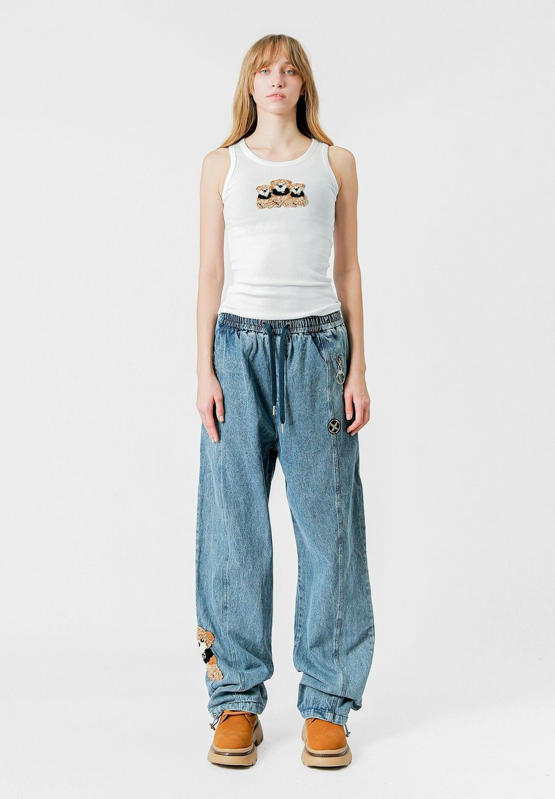 Camiseta sin mangas blanca con diseño de oso, combinada con pantalones de mezclilla sueltos de color azul claro con acentos bordados y calzado marrón.