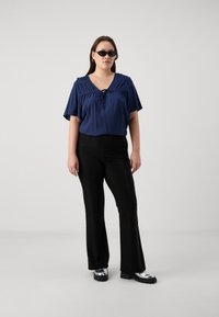 Blouse à manches courtes bleu marine avec des détails plissés ; pantalon noir évasé ; chaussures à plateforme argentées brillantes ; mannequin portant des lunettes de soleil noires.