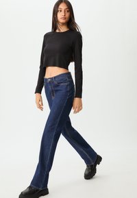 Crna skraćena majica s dugim rukavima u kombinaciji s plavim trapericama visokog struka. Traperice imaju ravne nogavice, vidljivo šivanje i klasičan dizajn.