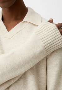 Gros plan sur une personne portant un pull en maille de couleur crème avec un col large et des poignets côtelés, la main reposant sur l'épaule opposée.