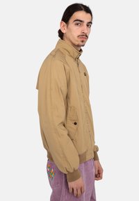 Tan bomber jacket with a high collar, front zipper, and side pocket. Textured fabric and elastic cuffs, worn with purple pants featuring colorful embroidery.