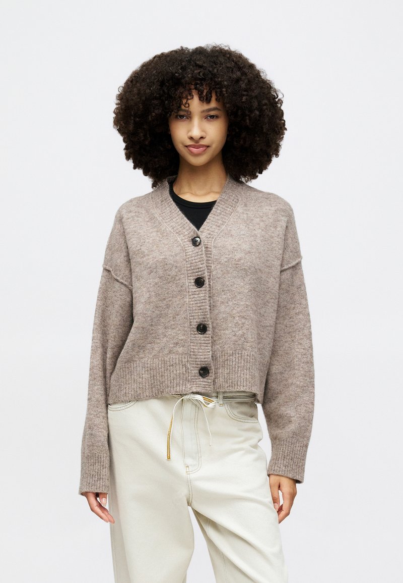 Ruda vilna kardiganas su V formos iškirpte, įglebtais rankogaliais ir ilgomis rankovėmis. Priekyje yra penki juodi mygtukai ir laisvas, trumpintas siluetas.