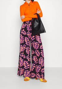 Haut zippé orange avec col, associé à un pantalon large noir orné de motifs de feuilles oranges et violettes. Complété par un sac noir.
