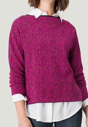 Femme portant un pull à motifs rose vif et noir par-dessus une chemise blanche à boutons, avec un jean bleu, recadrée au niveau de la taille.