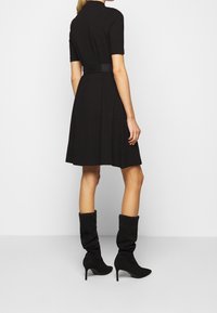 Schwarzes kurzes Kleid mit kurzen Ärmeln, tailliertem Oberteil und schwingendem Rock, aus strukturiertem Stoff und mit einem schwarzen elastischen Taillengürtel. Getragen mit schwarzen Kniestrümpfen.