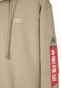 Haut à capuche beige en tissu doux, avec un patch logo sur le devant et une poche à glissière sur la manche avec une étiquette rouge "Remove Before Flight".
