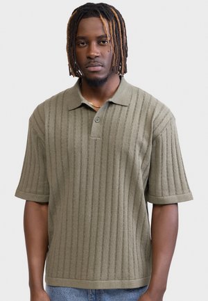 Homme avec des dreadlocks portant un polo en maille à manches courtes couleur vert olive avec des rayures verticales, debout devant un fond blanc uni.