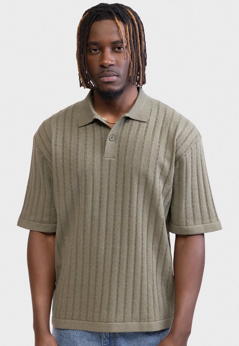Homme avec des dreadlocks portant un polo en maille à manches courtes couleur vert olive avec des rayures verticales, debout devant un fond blanc uni.