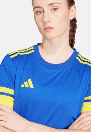 Giovane donna con capelli intrecciati, indossa una maglietta sportiva blu con strisce e logo gialli, braccia incrociate, espressione neutra, sfondo bianco.