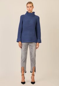 Fetter blauer Strickpullover mit hohem Kragen, gerippter Textur und umgeschlagenen Ärmeln, kombiniert mit grauen, verkürzten Jeans mit fransigem Saum.