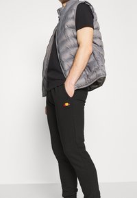 Grå quiltad pufferväst med dragkedja över en svart t-shirt; svarta sweatpants med en liten, färgad logotyp på fickan.