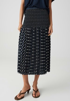 Femme portant une jupe midi noire à pois blancs et des sandales marron à talons, debout devant un fond clair uni.