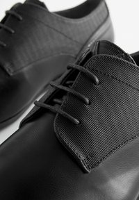 Zapatos negros con un cuerpo de cuero liso y una parte superior de tela texturizada. Presentan cordones negros y una punta redondeada. Diseño simple sin marcas visibles.