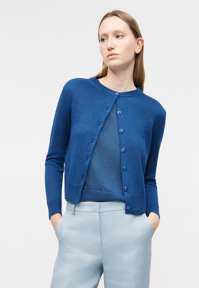 Γυναίκα που φοράει μπλε ζακέτα πάνω από μπλε τοπ και ανοιχτό γκρι παντελόνι, στέκεται με τα χέρια στις τσέπες μπροστά σε απλό φόντο.