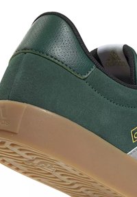 Baskets en daim vert foncé avec semelle en gomme, doublure de col noire et petit logo doré sur le côté près du talon.