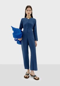 Blauwe linnen jumpsuit met lange mouwen en een gespleten voorkant, gecombineerd met een blauwe tote bag en zwarte sandalen met bontaccenten.
