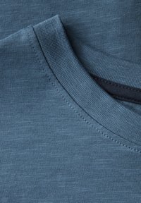 En närbild av ett blått bomullströja tyg, med rundad halsringning, fin textur och intrikata sömnadsdetaljer på kragen och fållen.