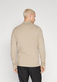 Beige színű, kötött, hosszú ujjú póló, textúrált felülettel, gallérral és testhezálló mandzsettákkal, hátulról nézve.
