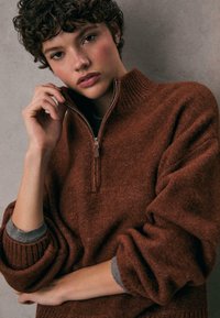 Brązowy sweter z dzianiny z wysokim kołnierzem i zamkiem błyskawicznym do połowy. Posiada luźne rękawy z szarymi akcentami. Miękka tekstura i swobodny design.