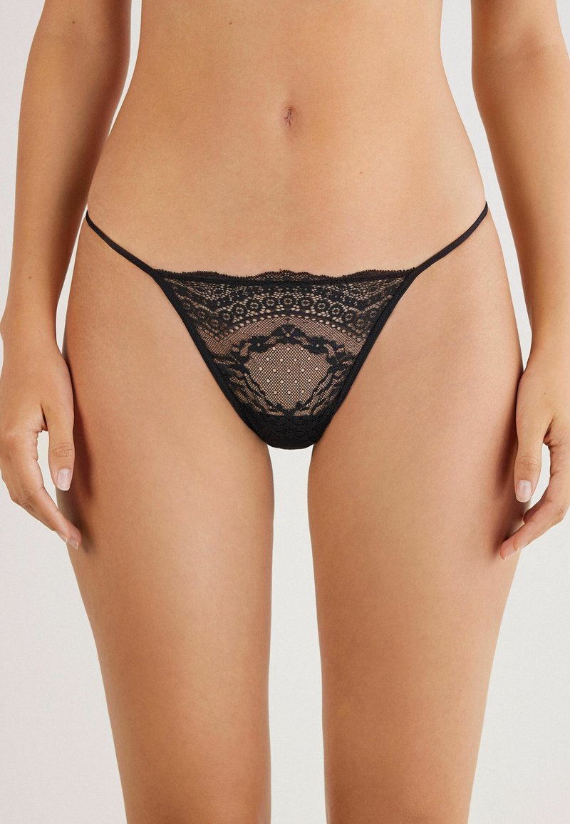 String en dentelle noire présentant des motifs complexes, une texture transparente et des fines bretelles, mettant en avant un design délicat et léger.