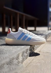 Grå lädersneaker med blå och vita ränder, rund tå, texturerad sula och en röd hälaccents. Lätt gummisula med en genomskinlig yta.