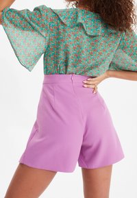 Blusa trasparente verde con stampa floreale e scollo arricciato, abbinata a pantaloncini lavanda a vita alta con cerniera e dettagli con tasche.