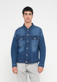 Junger Mann, der eine blaue Jeansjacke mit aufgesetzten Taschen und hellgraue Jeans trägt, steht vor einem schlichten weißen Hintergrund.