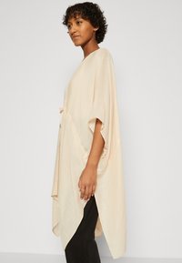 Beige, løs-fittende poncho med korte ermer, teksturert stoff, buet kant og justerbar snor i midjen, båret over mørke bukser.
