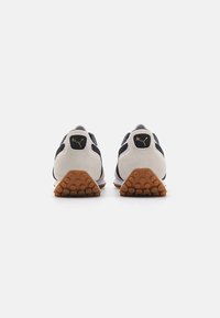 Zapatillas deportivas en blanco y negro con materiales de ante y textil. Suela de goma con un patrón de agarre texturizado. Logo dorado en el talón.