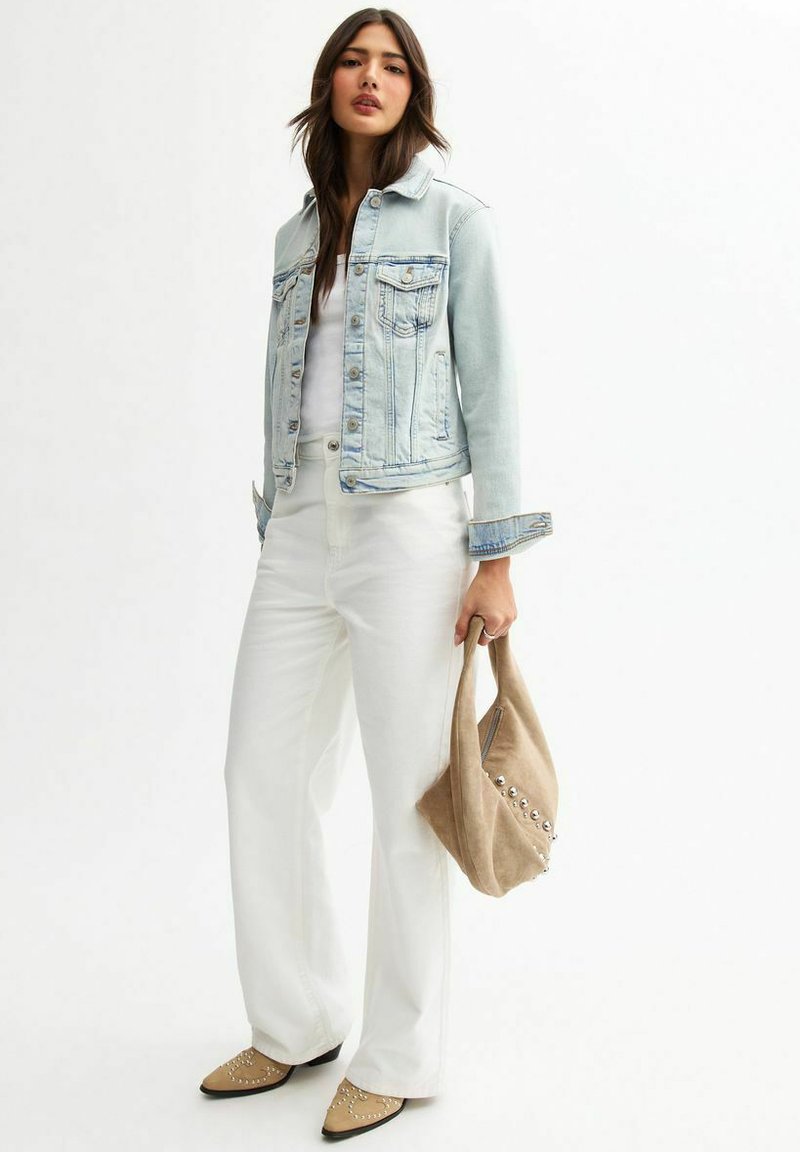 Lichtblauwe denimjack met twee voorzakken, witte fitted top, witte wijde broek en een tan tas met studs.