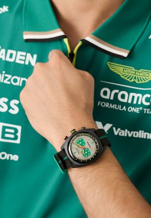 Melns hronogrāfa pulkstenis ar zaļiem akcentiem, kuram ir pelēks ciparnīca un trīs pulksteņa skaitītāji. Nēsāts uz rokas, blakus zaļai polo kreklam.