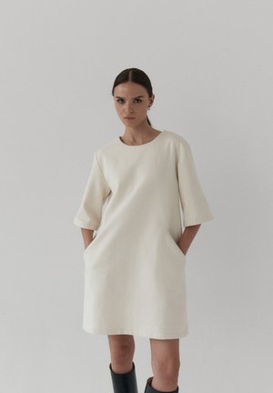 Kobieta o ciemnych włosach, ubrana w luźną, kremową sukienkę sięgającą kolan z kieszeniami oraz czarne kozaki sięgające za kolano na jednolitym jasnym tle.