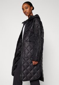 Black quilted puffer coat with a hood, featuring a full zipper closure, side pockets, and a subtle logo patch on the sleeve.
