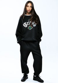 Sudadera negra de gran tamaño con el texto y logo en blanco "Grilly"; combinada con joggers negros ajustados y zapatillas negras. Textura de tela suave.
