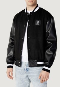 Svart varsityjacka med suédetyg på kroppen och läderärmar. Har vit och svart randig ribbad mudd och en logopatch på bröstet.