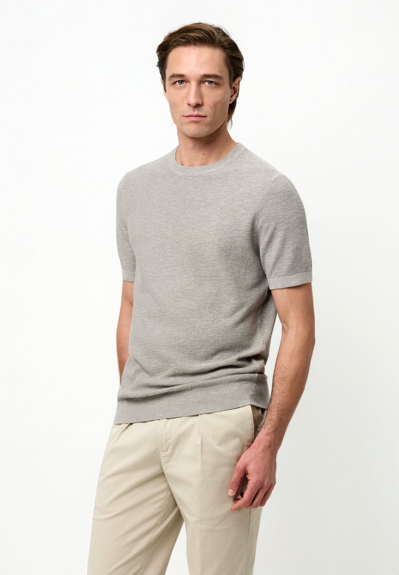 Homme portant un t-shirt en maille gris clair à manches courtes et un pantalon beige, debout avec une expression neutre sur un fond blanc.