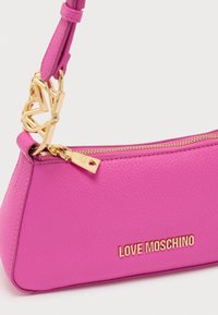 Ροζ δερμάτινη τσάντα χειρός με υφή, που διαθέτει χρυσό φερμουάρ, λογότυπο και διακοσμητική αλυσίδα από χρυσό μέταλλο στη λαβή.