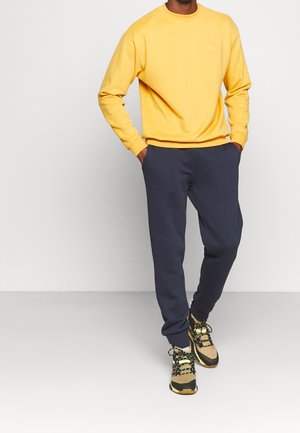 Sudadera amarilla de manga larga y cuello redondo, combinada con pantalones de chándal azul marino y zapatillas multicolor con acentos estampados.